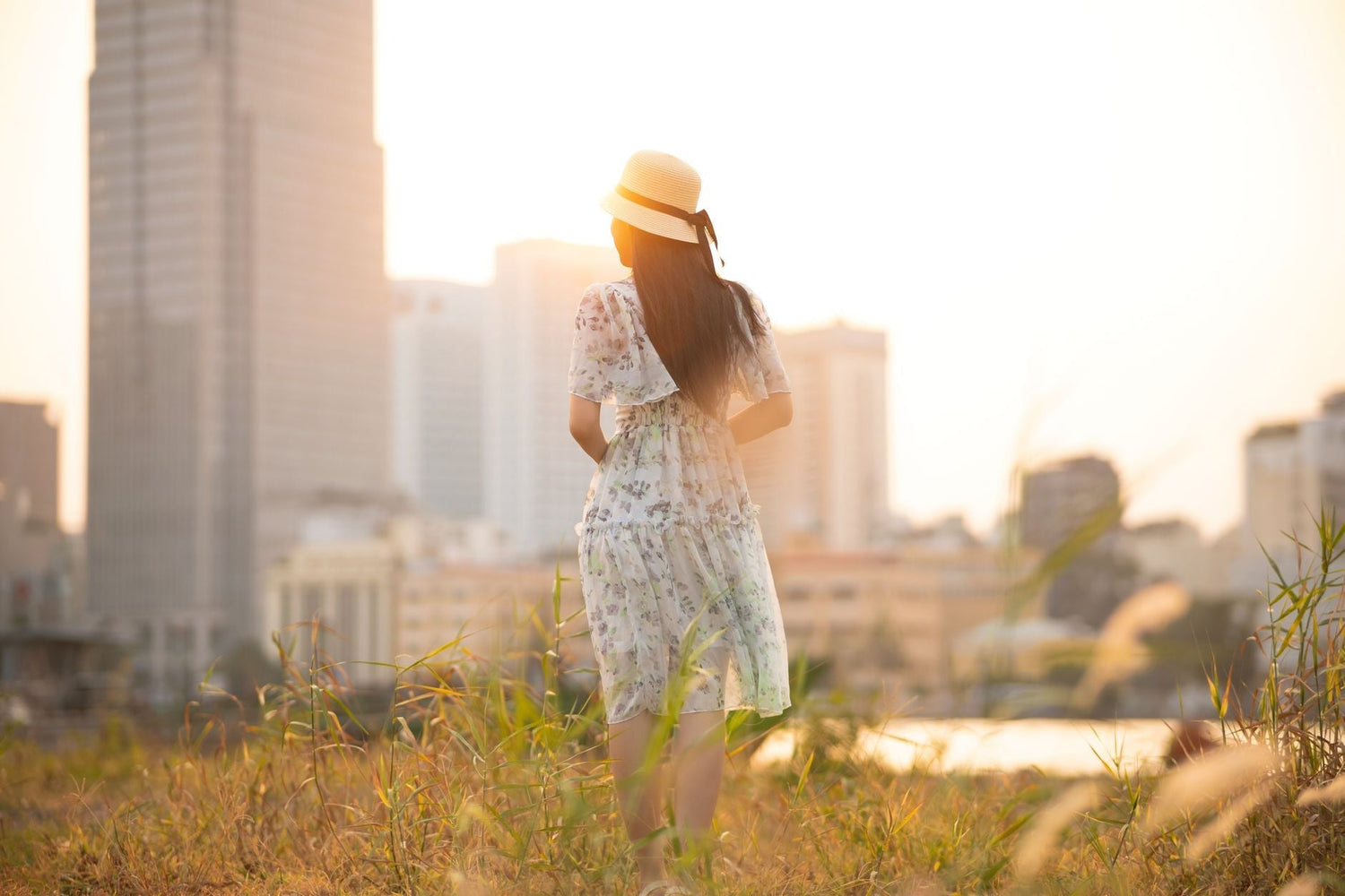 A woman in a summer dress and hat walks in a meadow with a city skyline in the background during daylight.