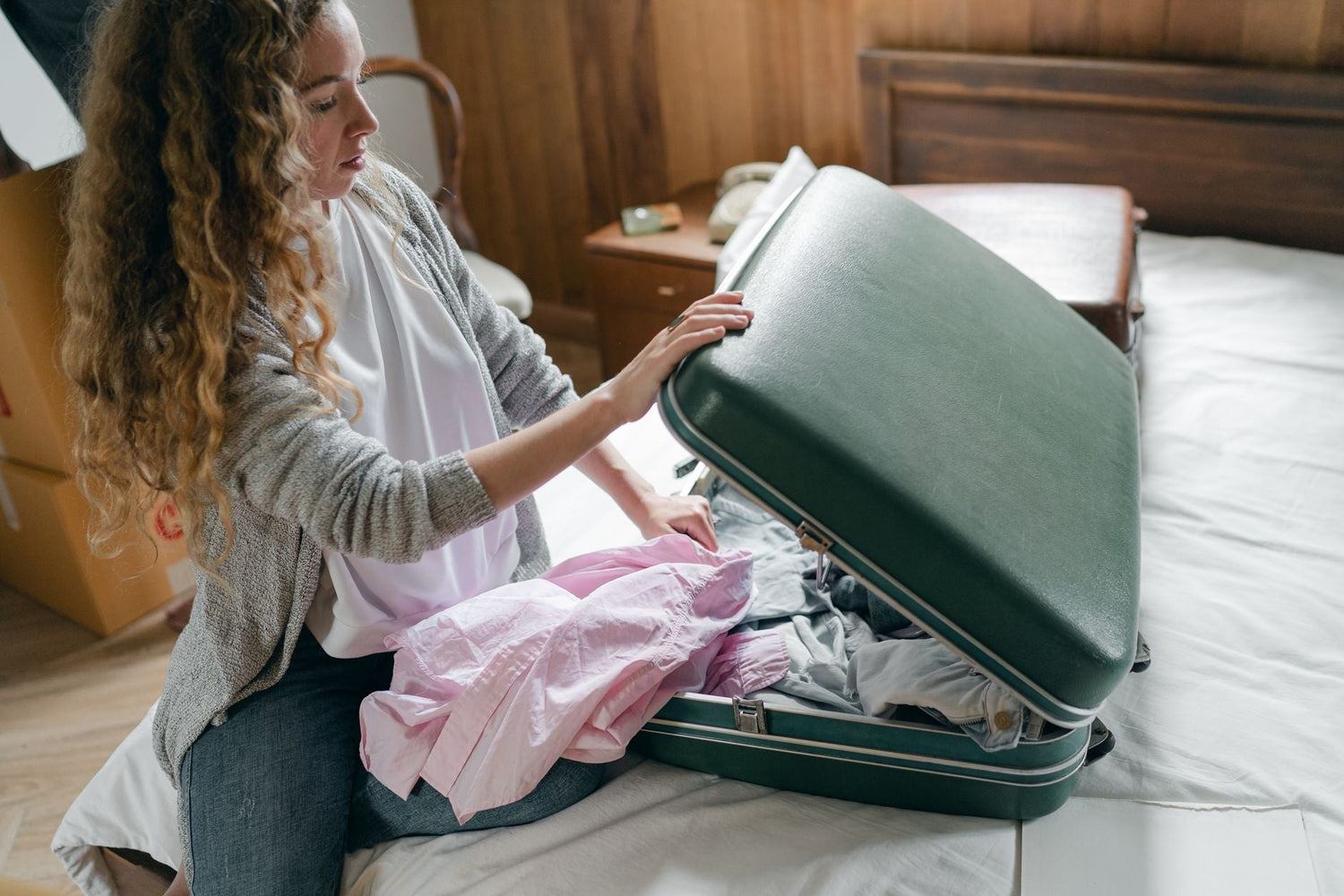 From above concentrated young female in casual wear with long curly hair sitting on bed and packing suitcase before trip